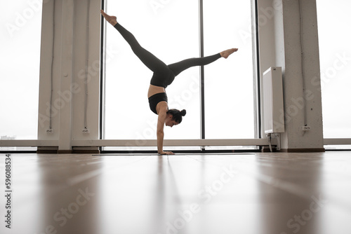 Young woman training alone at fitness studio with amazing city view throught panoramic window. Athletic female in black pants and top practicing headstand pose with widely spread legs on yoga mat.