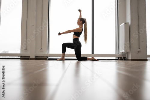 Strong caucasian woman standing with one knee on yoga mat and using dumbbells for arms exercises. Well-trained lady in black activewear having workout near panoramic windows with city view.