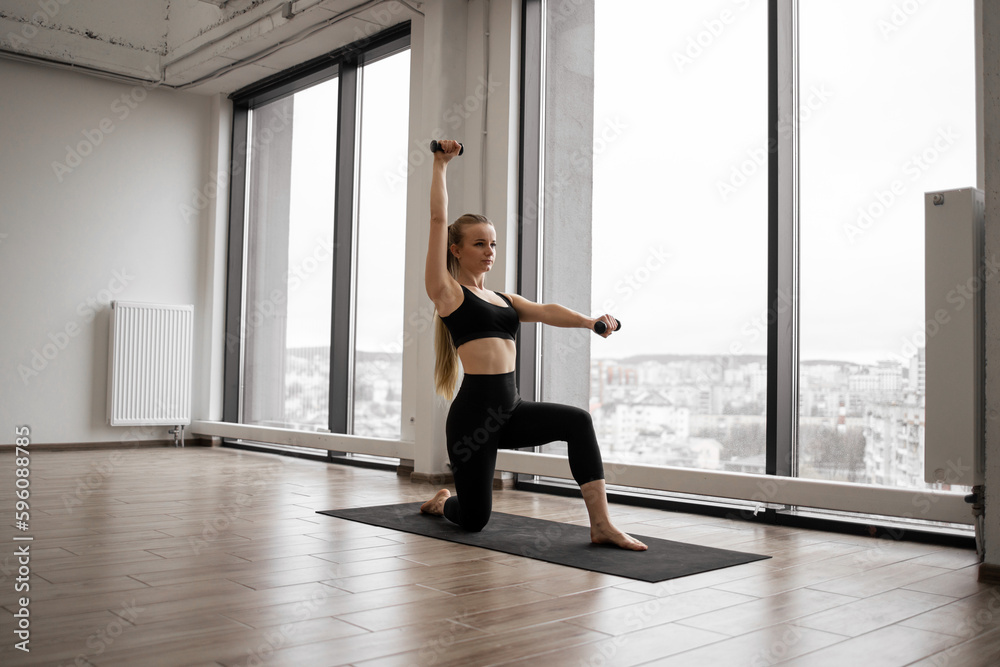 Fototapeta premium Strong caucasian woman standing with one knee on yoga mat and using dumbbells for arms exercises. Well-trained lady in black activewear having workout near panoramic windows with city view.