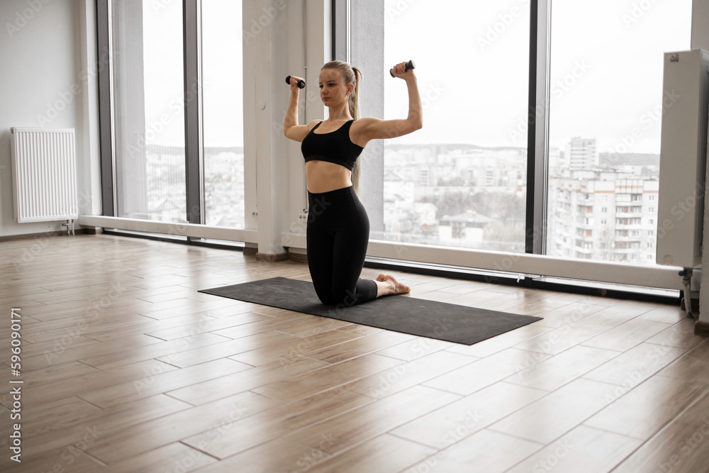 Front view of sporty young lady standing on knees on yoga mat and training arms with dumbbells. Caucasian fitness woman dressed in comfy active clothes performs sports exercises in studio. Front view of sporty young lady standing on knees on yoga mat and training arms with dumbbells. Caucasian fitness woman dressed in comfy active clothes performs sports exercises in studio.
