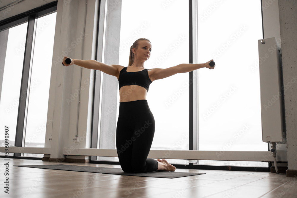 Front view of sporty young lady standing on knees on yoga mat and training arms with dumbbells. Caucasian fitness woman dressed in comfy active clothes performs sports exercises in studio.