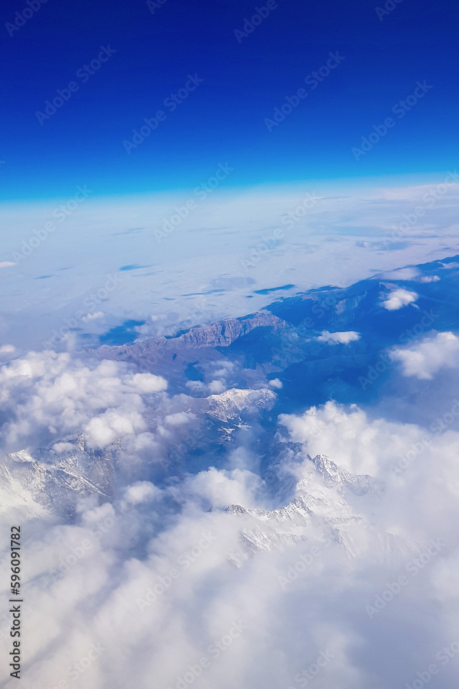 Fototapeta premium View from window of plane to blue sky and snowy mountain peaks in white clouds. High quality photo
