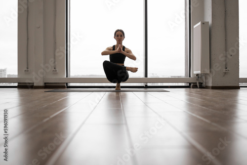Strong active lady balancing on one leg and keeping hands in namaste gesture at training room. Caucasian woman in sport clothes performing advanced yoga pose with french windows on background.
