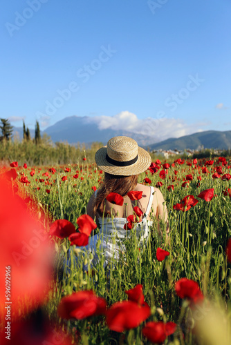 Young woman wearing white cotton dress and straw hat in the middle of red poppies field on an environment friendly flower farm. Close up, copy space, mountains on the background.
