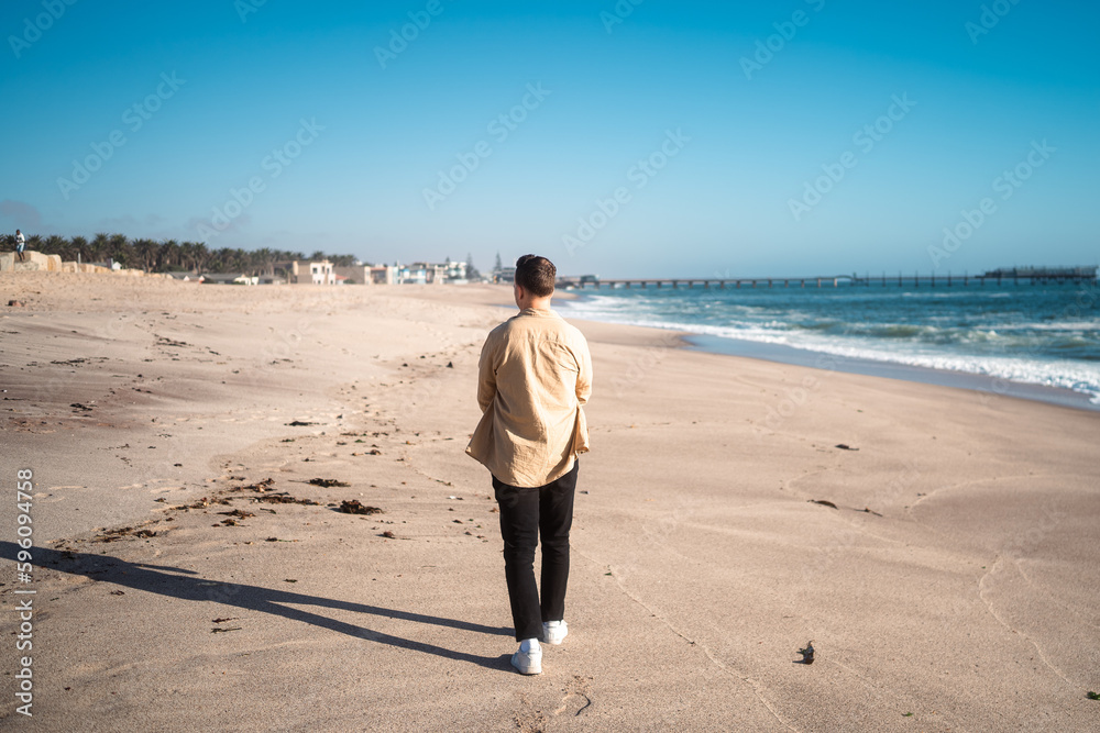Strolling along the shore: Young man enjoying a peaceful walk on the beach Strolling along the shore: Young man enjoying a peaceful walk on the beach