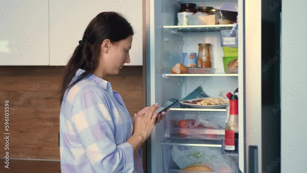 A woman is planning a food list near the refrigerator using a mobile app. A woman is planning a food list near the refrigerator using a mobile app.