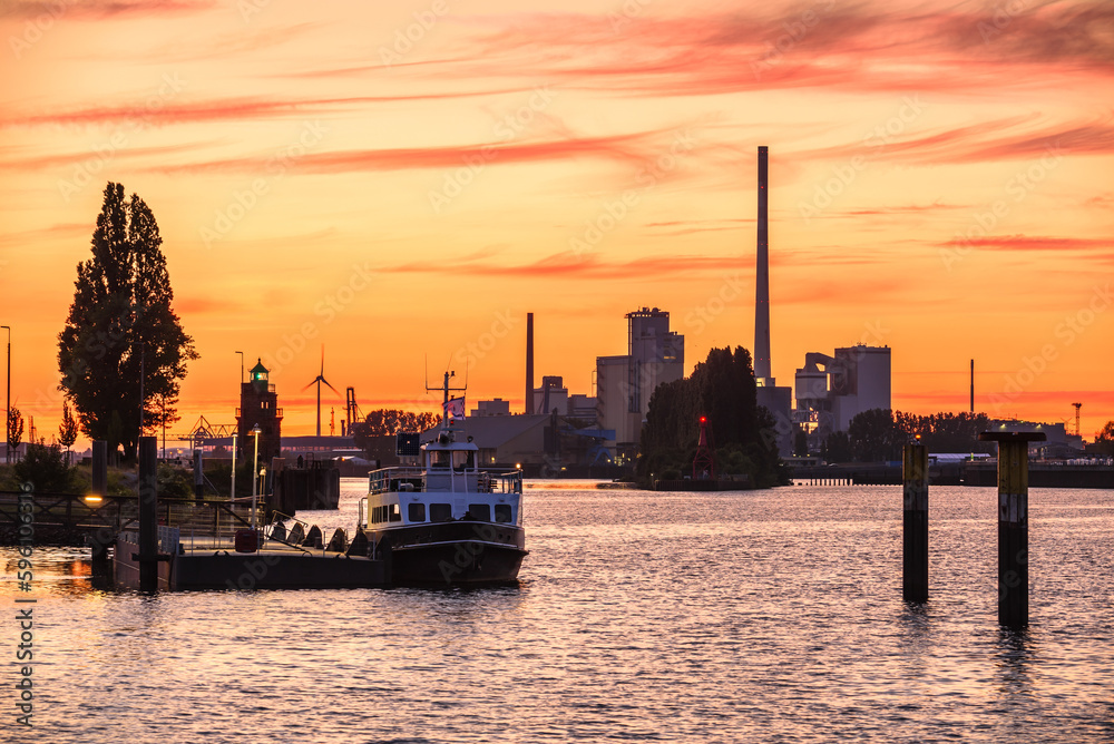 Fiery sunset sky over a river harbour. Industrial buildings, wind turbines and a lighthouse are in background.
