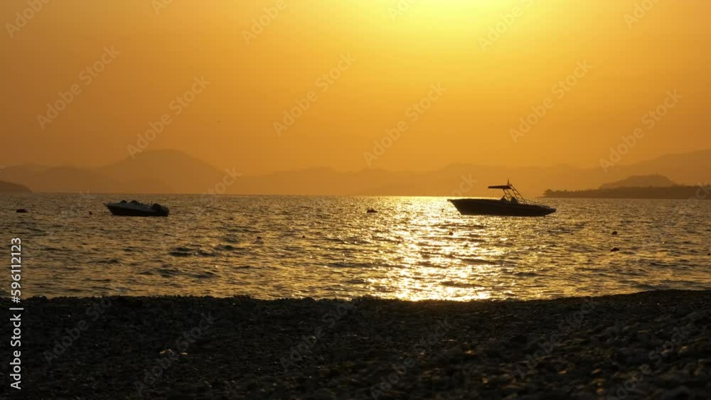 Moored boats silhouette during sunset. A view of floating boats silhouette by the pebbles beach during orange sunset in summer. Moored boats silhouette during sunset. A view of floating boats silhouette by the pebbles beach during orange sunset in summer.