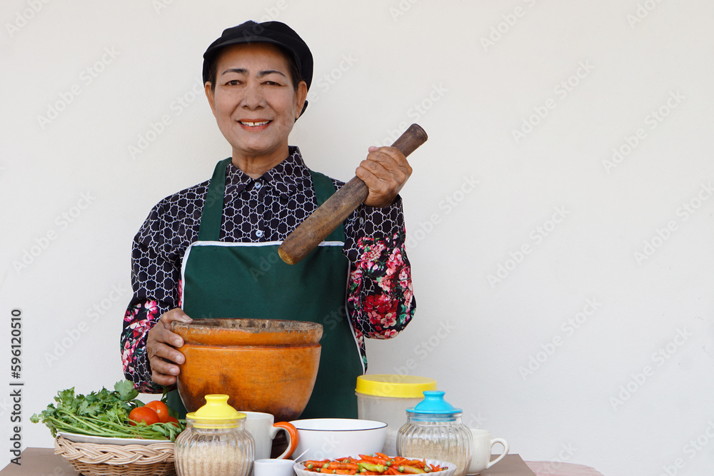 Φωτογραφία Happy Asian senior woman is cooking, wear chef cap and apron, holds pestle, mortar and plate of chillies