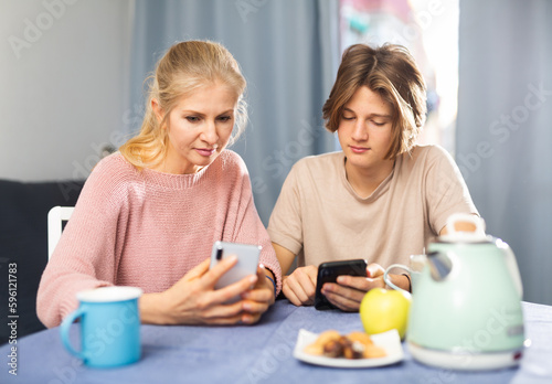 Portrait of mother and teenager son using phones during breakfast indoors