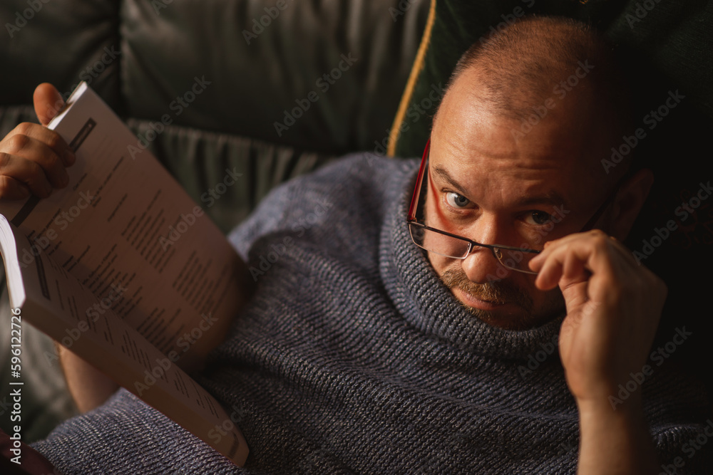 Fototapeta premium A man with glasses is reading a book while lying on the couch. Middle-aged man of ordinary appearance in casual clothes at home. Reading, learning.