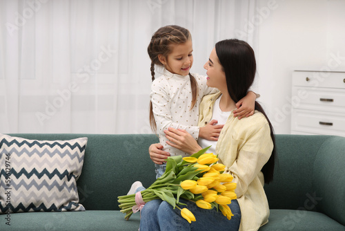 Happy woman with her daughter and bouquet of yellow tulips on sofa at home, space for text. Mother's day celebration
