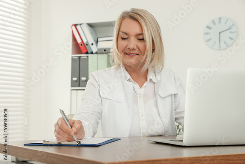 Doctor with laptop and clipboard writing at wooden table in clinic