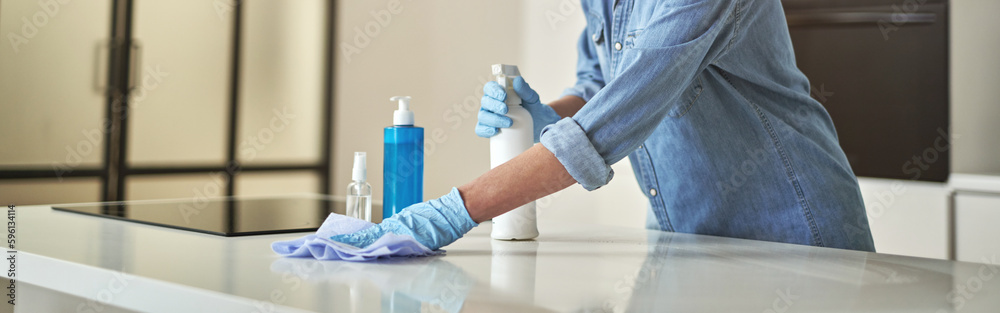 Happy mature woman in rubber gloves smiling at camera, using detergent spray and cloth while cleaning surface in the kitchen