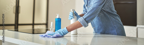 Happy mature woman in rubber gloves smiling at camera, using detergent spray and cloth while cleaning surface in the kitchen