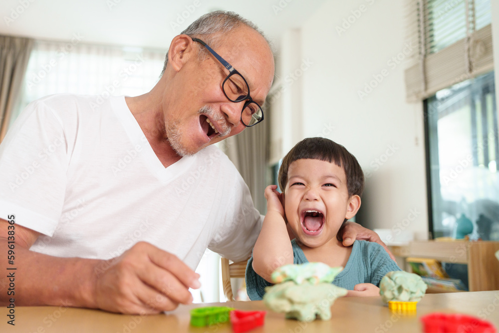 Portrait of Laughing Asian Grandfather and Grandson making toys from plasticine at home. Senior man and Little child boy playing dough. Creativity and hobby.