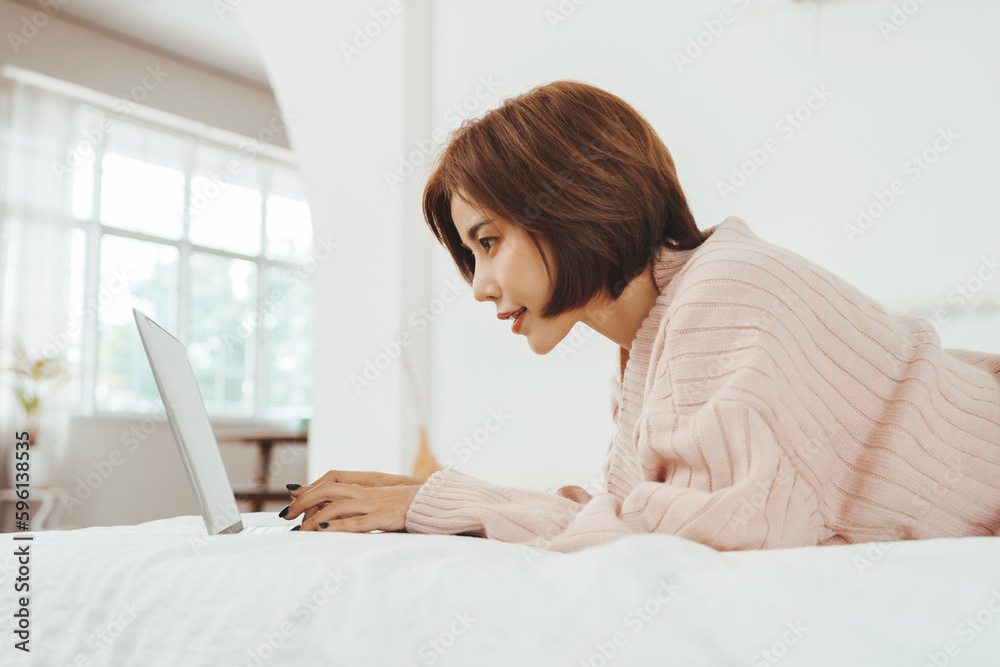 Young Beautiful Asian Korean woman lying on the bed and having online shopping on a laptop, Chatting and happy smiling at home Young Beautiful Asian Korean woman lying on the bed and having online shopping on a laptop, Chatting and happy smiling at home