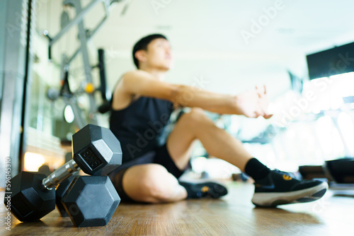 Happy cheerful Asian sportsman relaxing - sitting on the floor after done a weight training exercise in an indoor fitness or gym. Man resting after did a wight training workout.