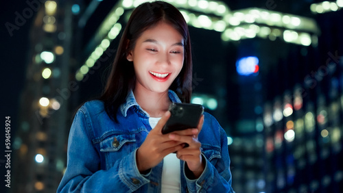 Beautiful young Asian woman walking in city street at night using mobile smartphone. Happy woman standing and typing messages on her mobile phone