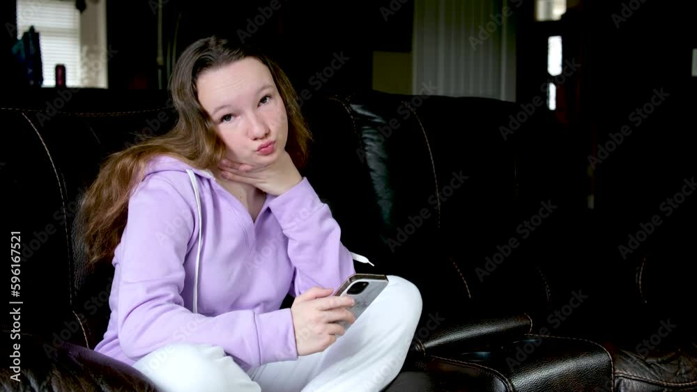 teenage girl sits on sofa in hands holding phone with hand holds throat looks into frame, makes eyes, shakes head, smiles, spends calm time In dark room, place for text teenage activities teenage girl sits on sofa in hands holding phone with hand holds throat looks into frame, makes eyes, shakes head, smiles, spends calm time In dark room, place for text teenage activities