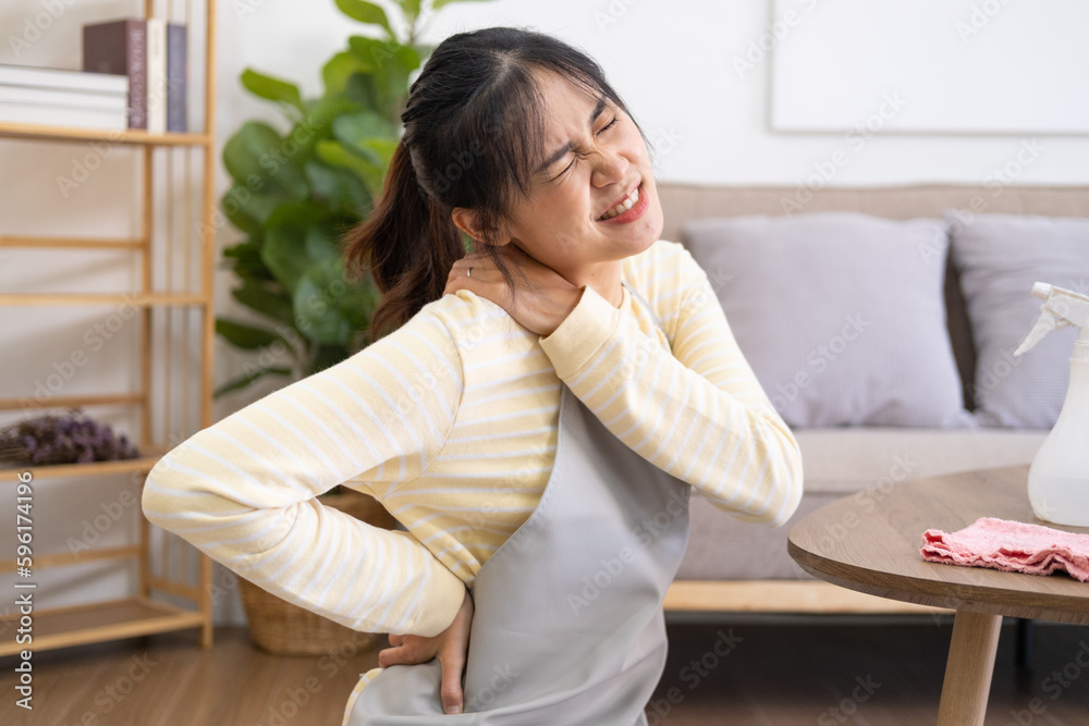 Asian woman doing housework wearing apron sitting on floors with shoulder feeling hurt painful in the living room. young lady Cleaning table by spraying Cleaning Products and wiping.. Asian woman doing housework wearing apron sitting on floors with shoulder feeling hurt painful in the living room. young lady Cleaning table by spraying Cleaning Products and wiping..