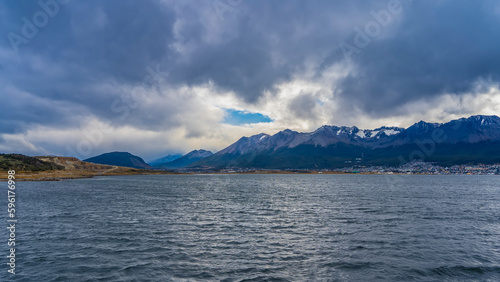 On the shore of the Beagle Canal, you can see city houses, a picturesque snow-capped mountain range. Through a gap in the gray low clouds, you can see the blue sky. Ripples on the water. Argentina.