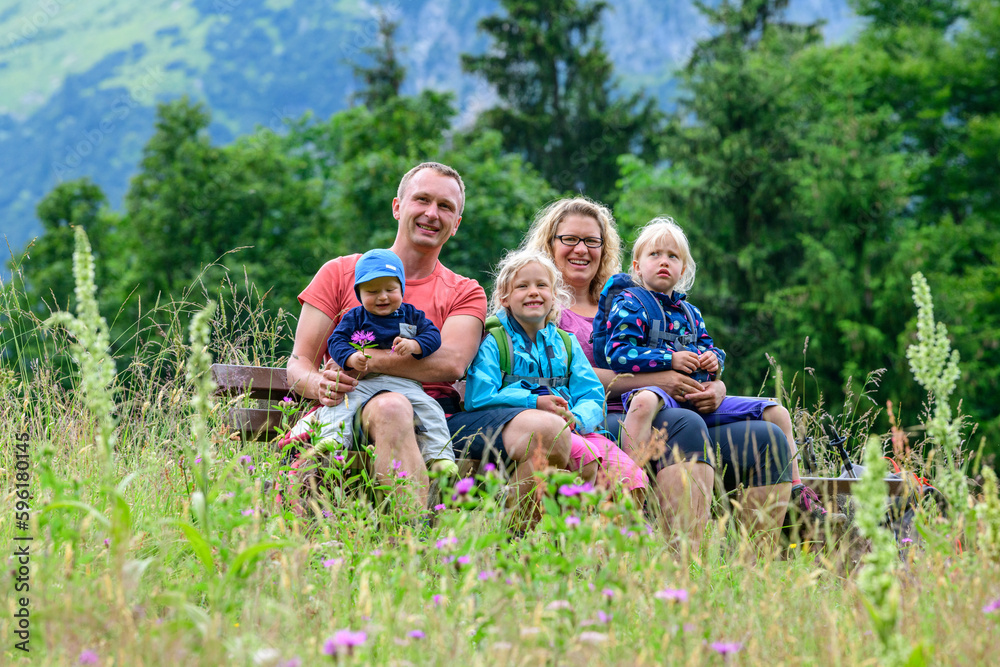 Obraz premium Fröhliche, junge Familie posiert auf einer Bank inmitten einer Wiese im Gebirge