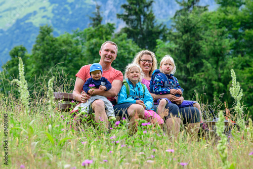 Fröhliche, junge Familie posiert auf einer Bank inmitten einer Wiese im Gebirge © ARochau