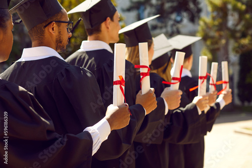 Group of diverse university students on graduation day. Happy mixed race multiethnic college graduates in black caps and gowns holding diplomas in hands while standing in row at outdoor ceremony
