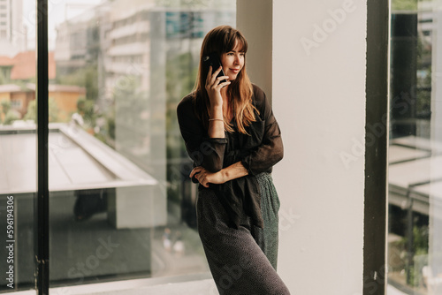 Stylish business woman in fashionable casual outfit talking by mobyle phone during work day in modern business center.