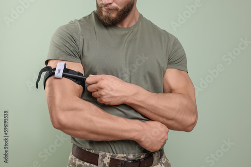 Soldier in military uniform applying medical tourniquet on arm against light olive background, closeup