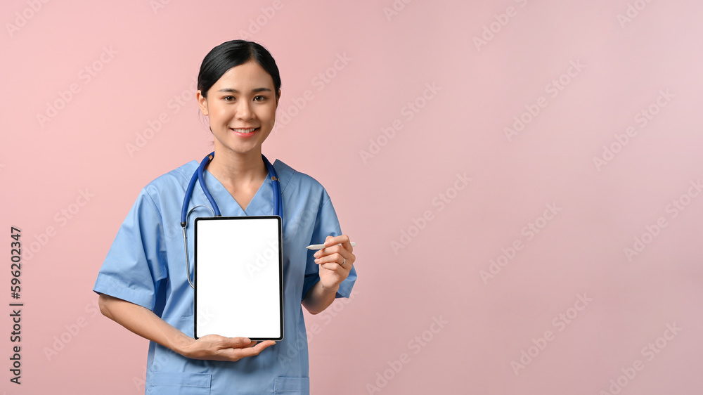 Happy doctor showing blank screen tablet to the camera while standing on pink isolated background.