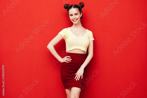Young beautiful smiling female in trendy summer yellow t-shirt and skirt. Carefree woman with two horns hairstyle posing near red wall in studio. Positive model having fun. Cheerful and happy