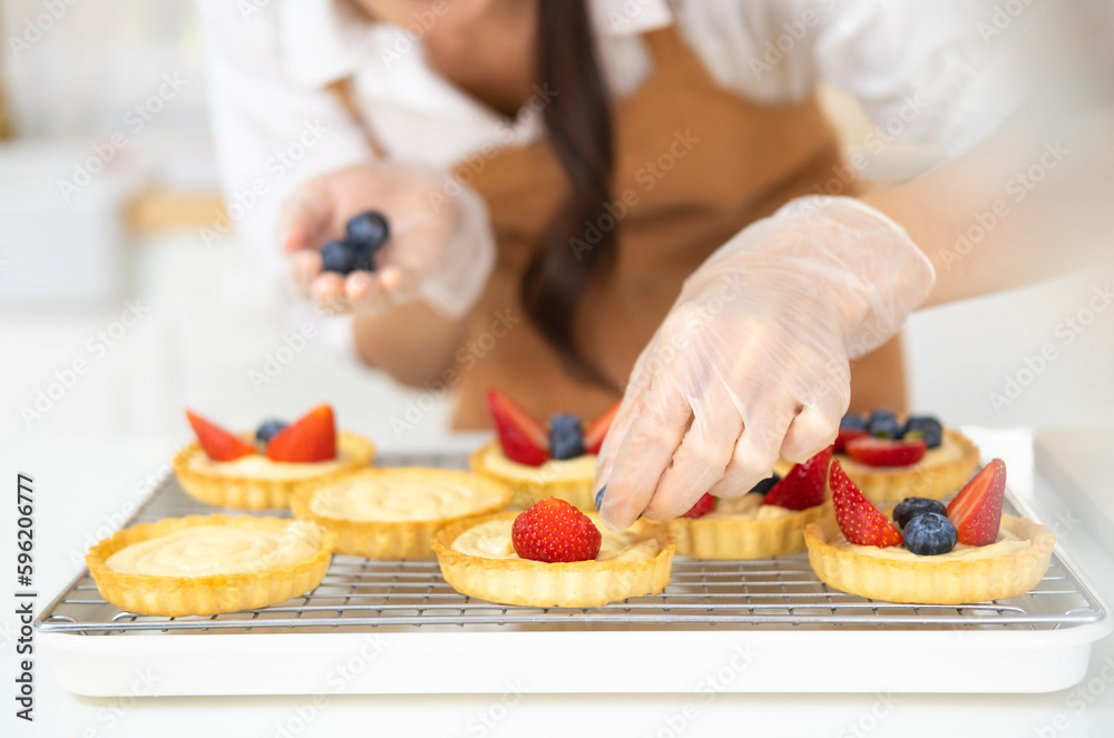 Young beautiful woman is baking in kitchen , bakery and coffee shop business. Hands in cooking gloves decorating freshly baked tart with blueberries strawberry fresh fruit. homemade bakery at home.