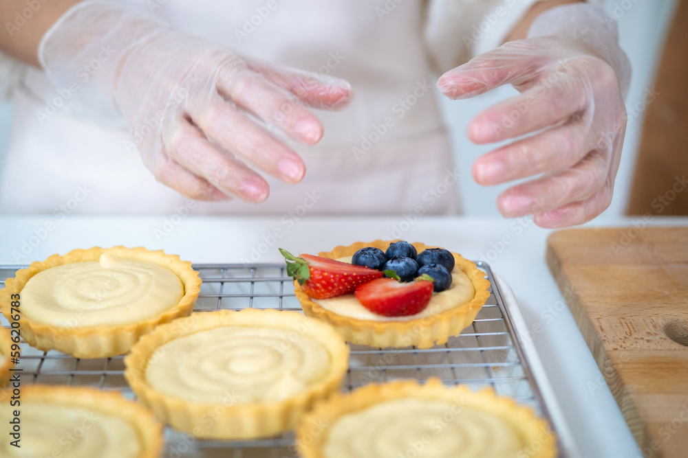 close up of hands in cooking gloves Baker adding blueberries strawberry fresh fruit to a tart on white table in Kitchen. housewife baker wear apron making fruit tart. homemade bakery at home.