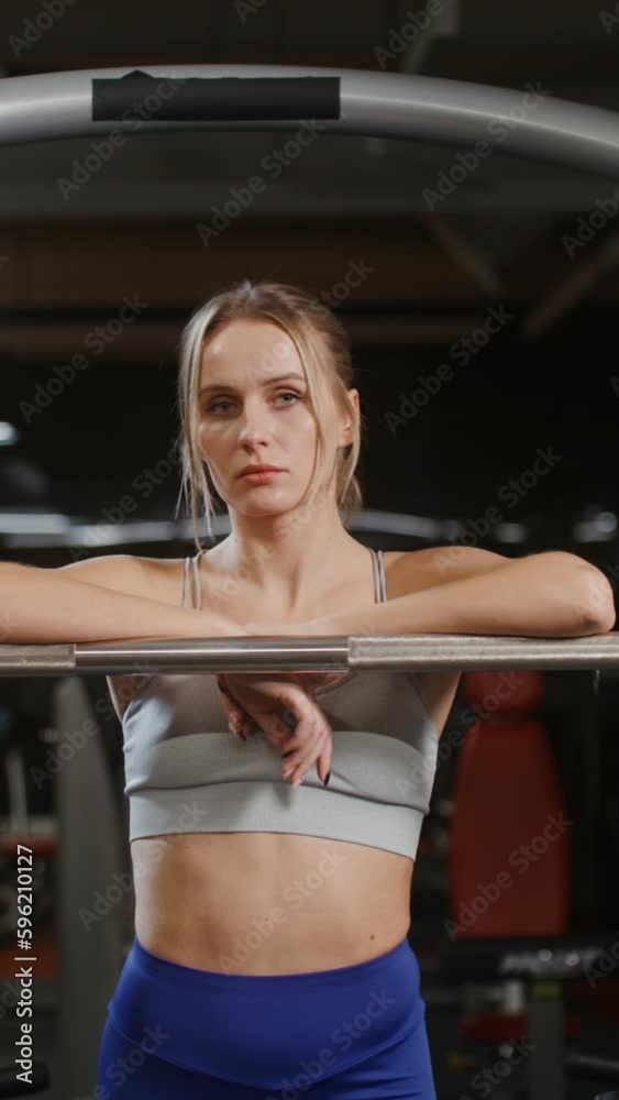A young blonde woman of European appearance, dressed in a sports uniform, looks at the camera, leaning on an exercise machine, standing in the gym alone A young blonde woman of European appearance, dressed in a sports uniform, looks at the camera, leaning on an exercise machine, standing in the gym alone