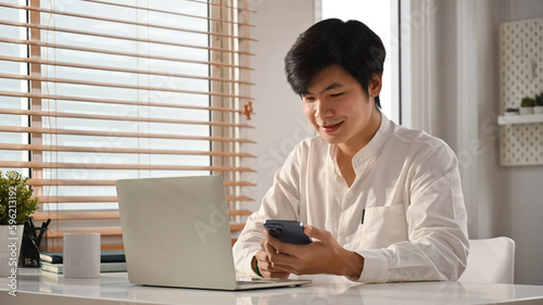 Handsome asian man entrepreneur sitting at workplace texting messages, chatting in social on smartphone