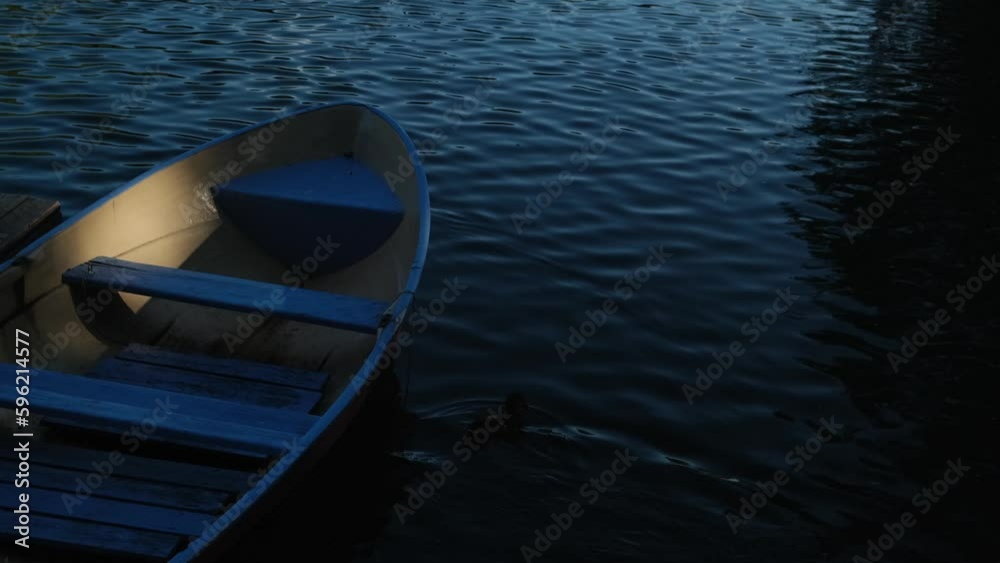Old wooden boat moored to the shore of quiet river in evening light. Tranquil summer marine landscape. Empty fishing boat sways in peaceful lake water.