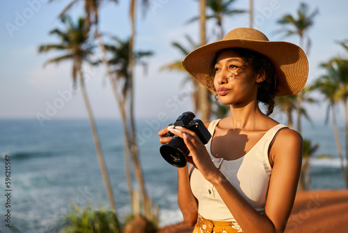 Fotografi Young african female model tourist with camera in colorful clothes and straw hat taking pictures at tropical location at sunrise