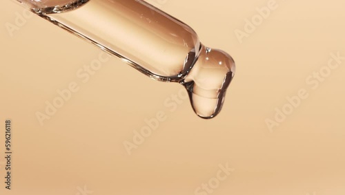 Essential oil dripping from a glass dropper, beige background, close-up. The concept of dermatology and cosmetology.