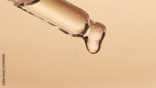 Essential oil dripping from a glass dropper, beige background, close-up. The concept of dermatology and cosmetology. photo