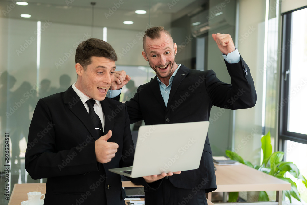 Two young businessmen looking at laptop smiling and expressing joy that the business succeeded according to their plans colleague talking to each other describing good partnership Two young businessmen looking at laptop smiling and expressing joy that the business succeeded according to their plans colleague talking to each other describing good partnership