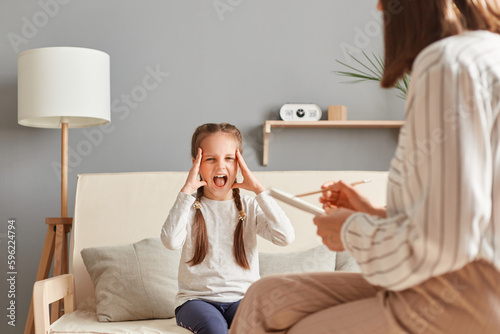 Despair hysterical child sitting on sofa screaming holding head having problems home violence mental mind disorders psychological therapy appointment.