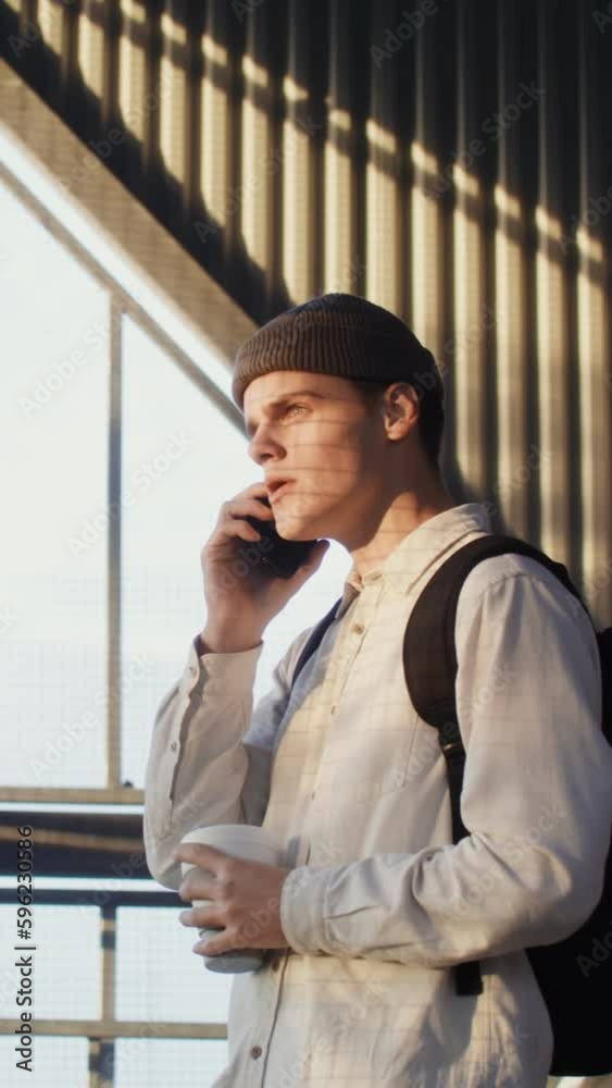 A young man of European appearance, in a knitted hat with a backpack on his back, is talking on the phone, laughing cheerfully