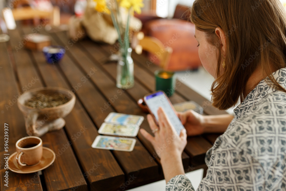 Fototapeta premium Beautiful young woman is guessing on cards with tarot, runes on wooden table and uses an online app in phone to interpret divination, home interior,candles and dry herbs..