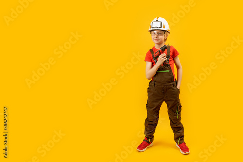 Boy as worker on yellow background. Child wears protective helmet and holds drill in his hand. Copy space, mock up