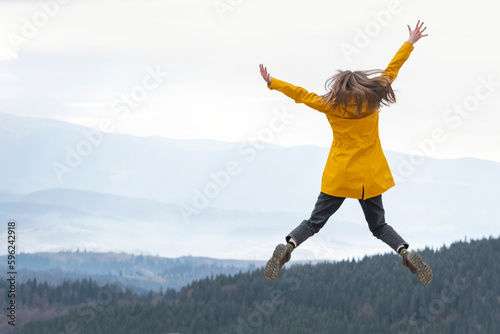 Φωτογραφία Happy girl in yellow jacket jumps with hands up to the sky on the top of mountains
