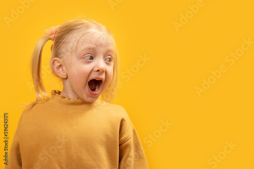 Surprised or frightened little girl with her mouth open looks to the side. Portrait of preschool girl on yellow background. Copy space.