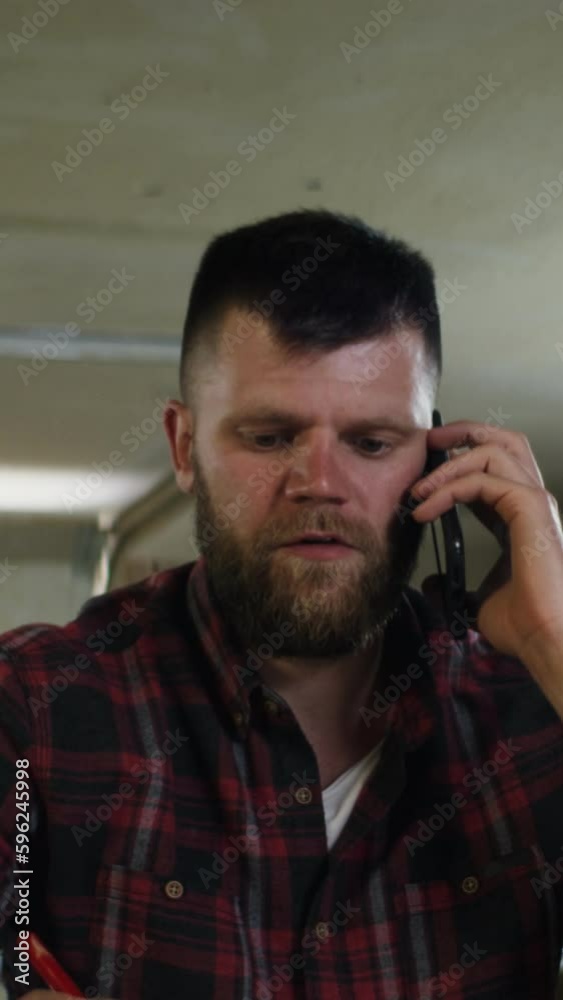 a serious man of European appearance with a beard, in a plaid shirt, talking on the phone, making notes in a notebook and looking into a laptop in a carpenter's shop, many wooden items are visible a serious man of European appearance with a beard, in a plaid shirt, talking on the phone, making notes in a notebook and looking into a laptop in a carpenter's shop, many wooden items are visible