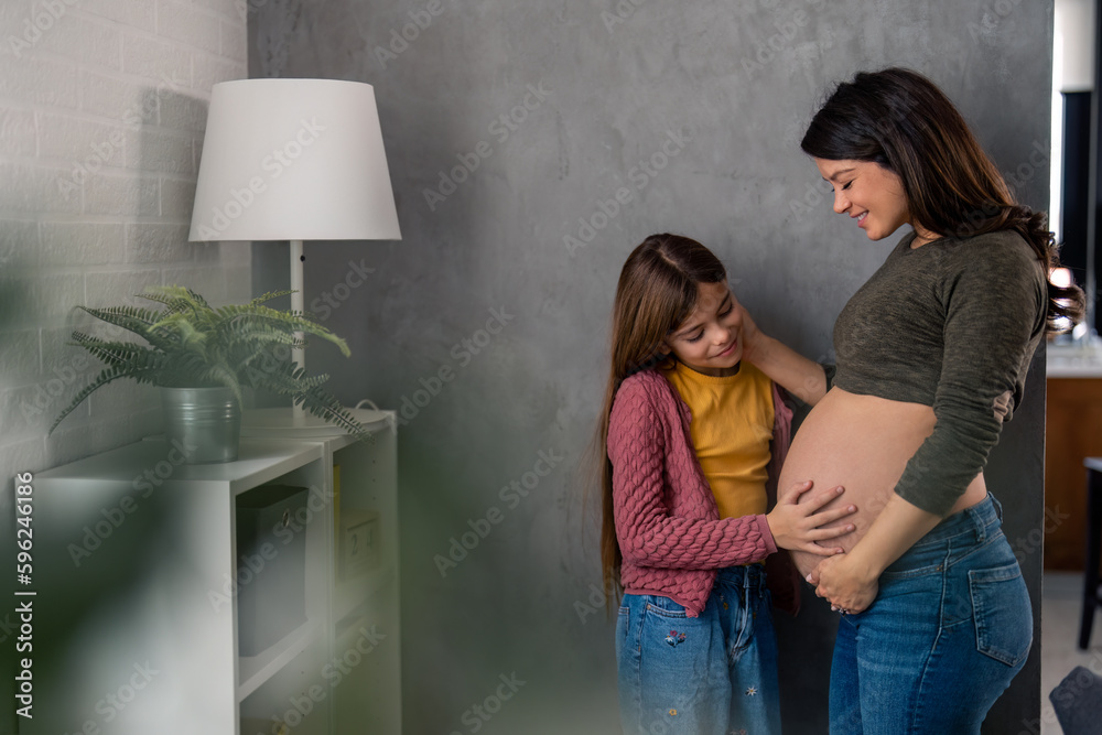 © Dorde - Photo of a pregnant woman with her daughter. Sweet little girl cuddling the belly of her caring mother during the day at home. Family, love and support.